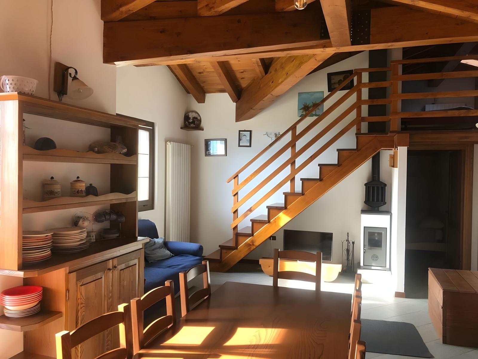 Wohnraum mit Holztreppe, Essbereich und Kaminofen. Holzmöbel und Regale sichtbar.
