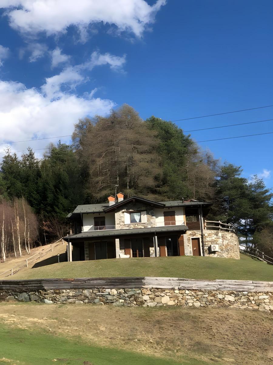 Haus mit Steinmauer und Terrasse auf grünem Hang unter blauem Himmel.