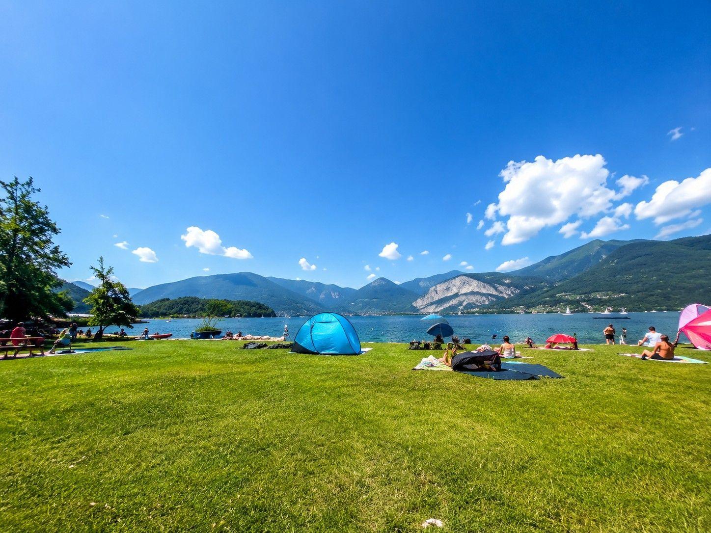 Grünes Gras am See mit Zelten und Sonnenschirmen. Berge im Hintergrund und blauer Himmel.