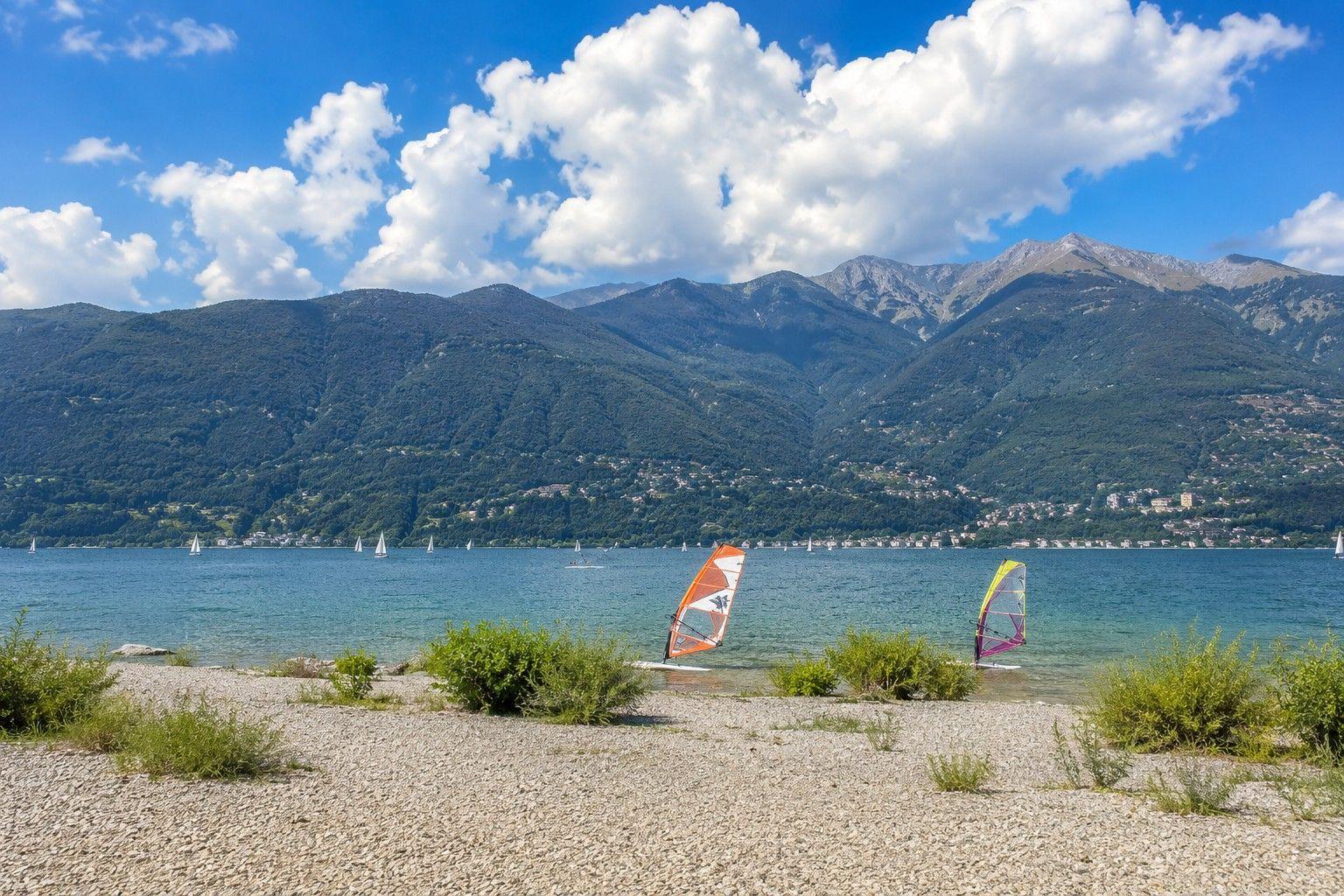 Strand mit Windsurfbrettern vor See und Bergen unter blauem Himmel mit Wolken.