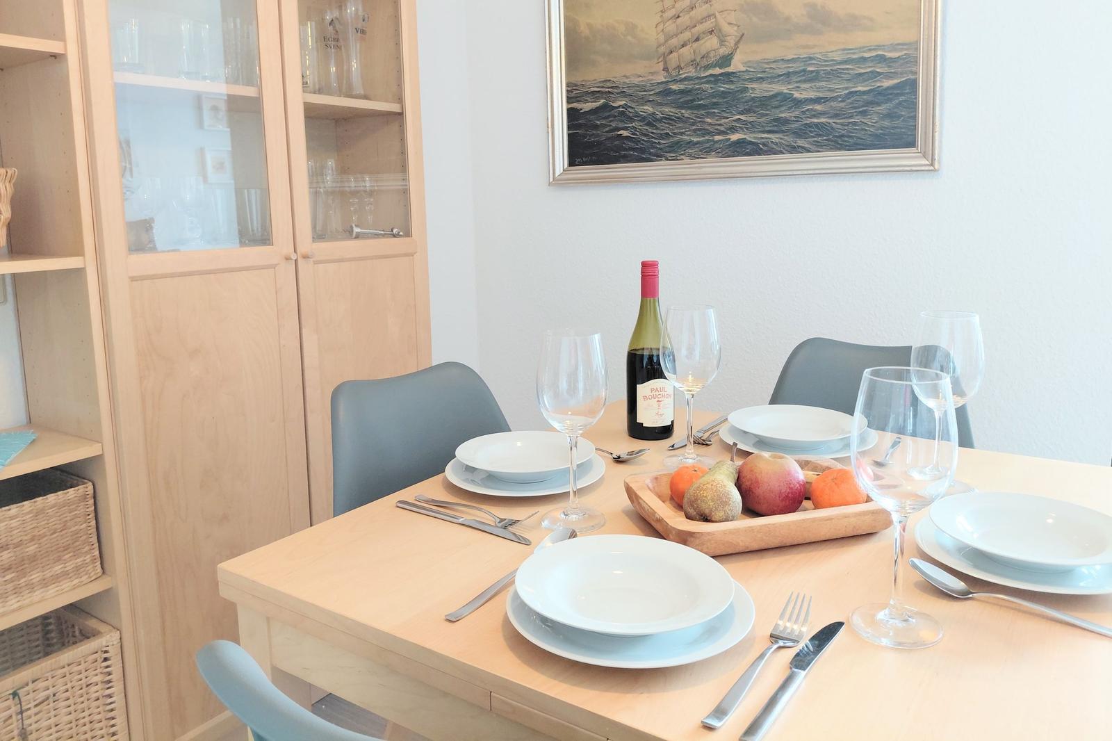 Esstisch mit Geschirr, Weinflasche und Obst. Hintergrund: Wandbild und Glaswand.