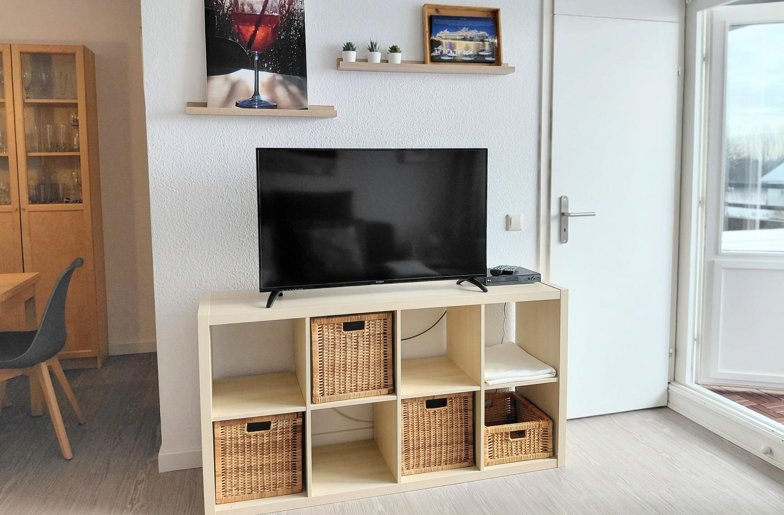 TV auf Holzregal mit Körben, Wandregal mit Bildern und Pflanzen, Tür und Fenster im Hintergrund.