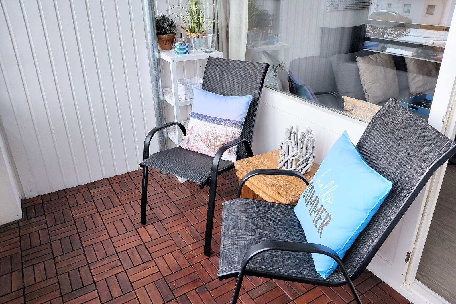 Terrasse mit zwei Stühlen, Kissen und Holztisch. Hintergrund zeigt Wohnraum.