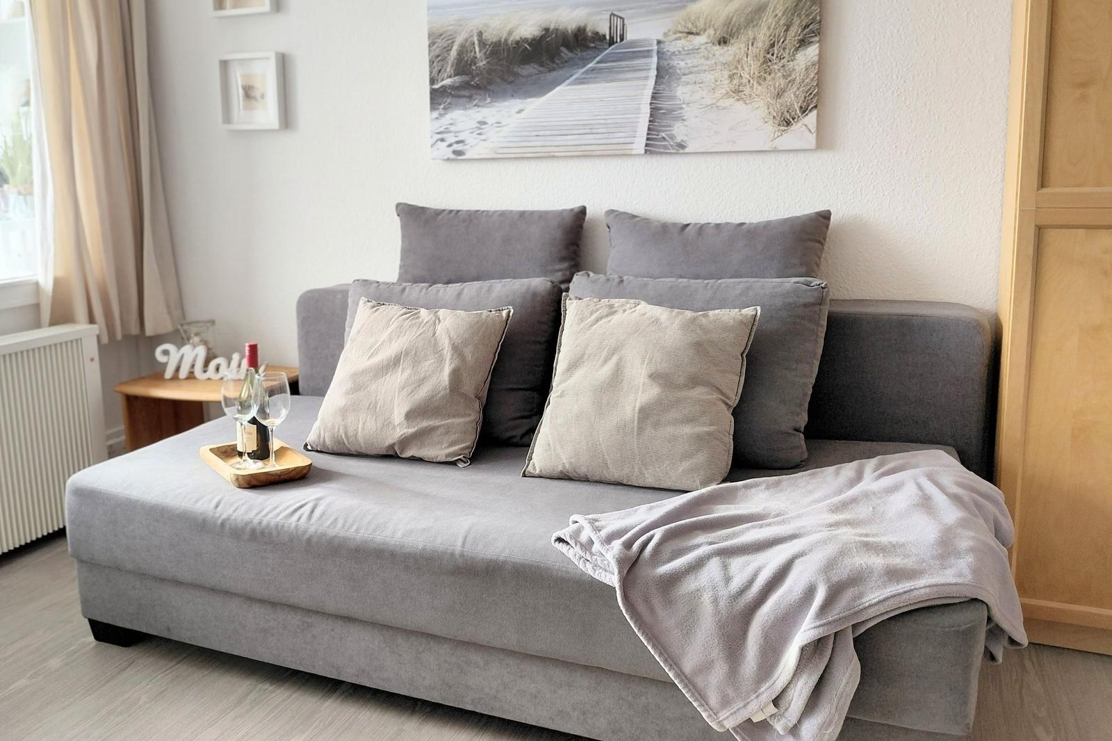 Graße Couch mit Kissen und Decke. Wandbild zeigt Strandweg. Nebenher kleiner Tisch mit Wein.