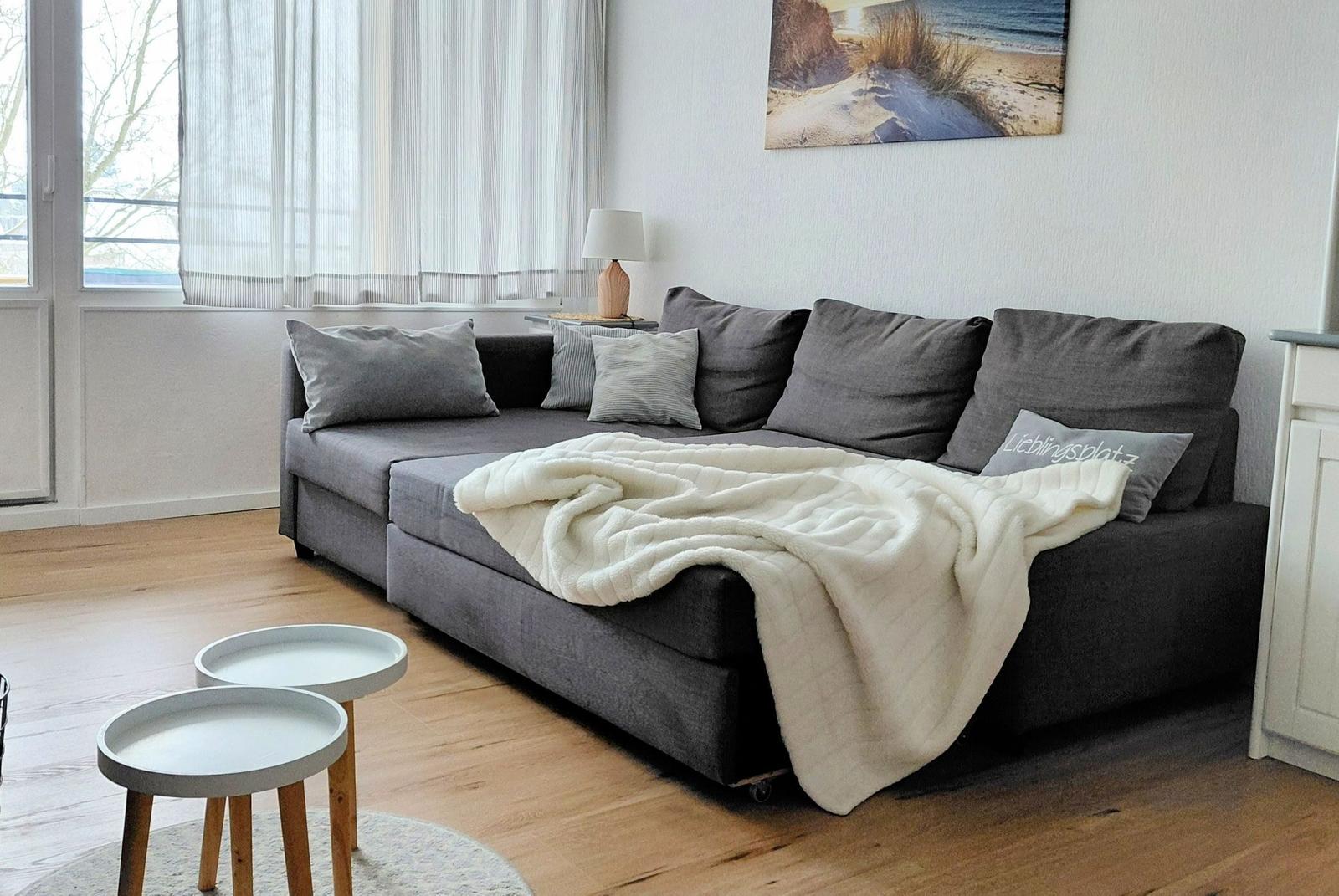 Ein grauer Sofa mit Kissen und einer Decke. Zwei runde Tische stehen auf dem Holzboden.