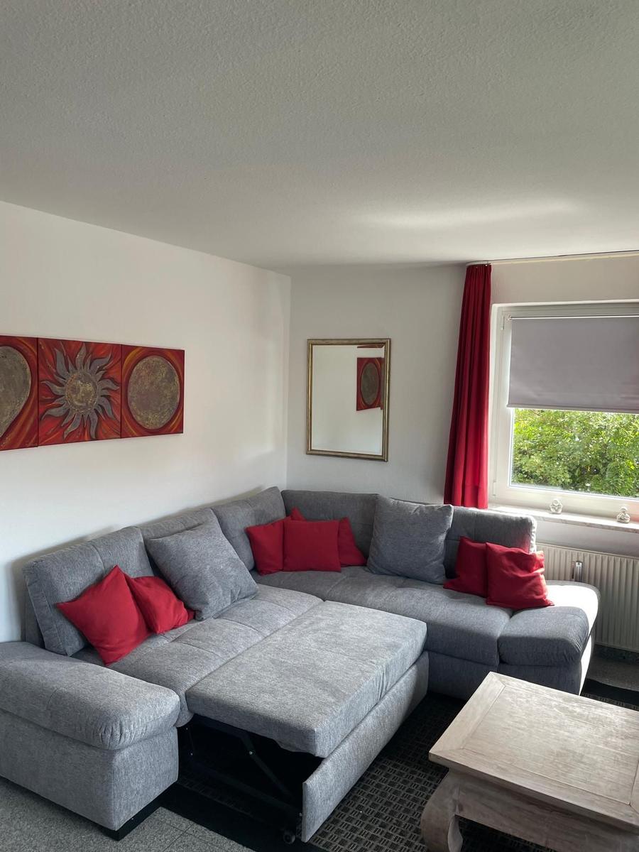 Ein Wohnzimmer mit grauem Sofa, roten Kissen und Wandbildern. Fenster mit rotem Vorhang.