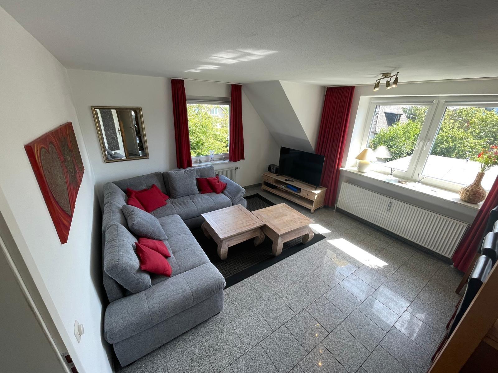 Wohnzimmer mit grauem Sofa, roten Kissen, Holztischen und großen Fenstern.