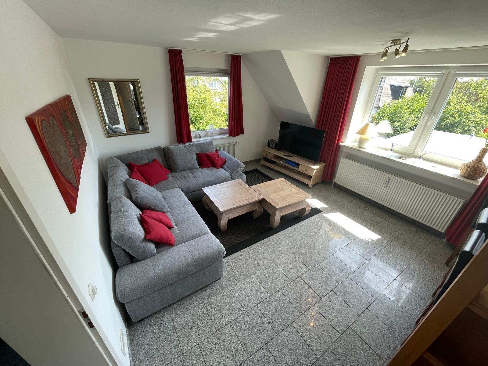 Wohnzimmer mit grauem Sofa, roten Kissen, Holztischen und Fenstern mit roten Vorhängen.