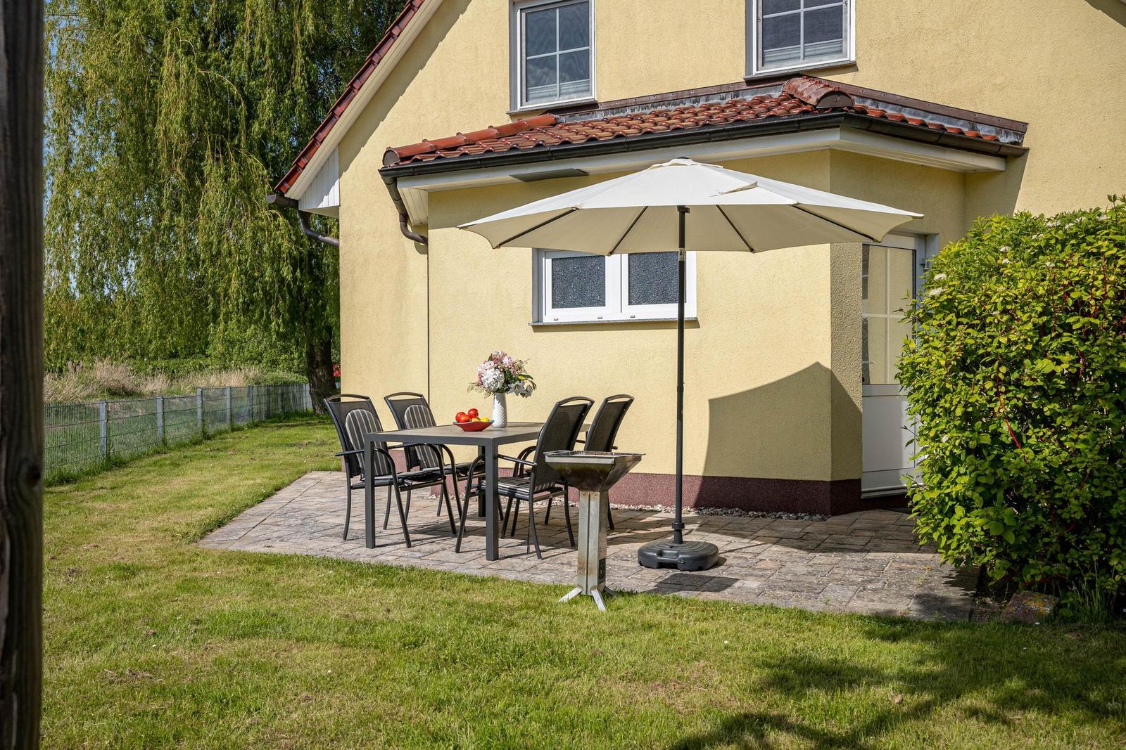 Terrasse mit Tisch, Stühlen, Sonnenschirm und Grill vor gelbem Haus mit Rasen und Bäumen.