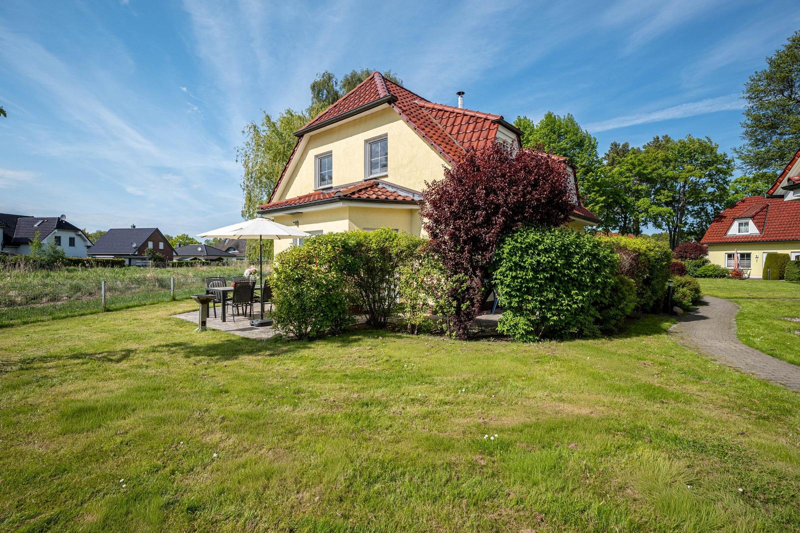 Ein gelbes Haus mit rotem Dach, umgeben von Gras und Büschen. Außentisch mit Sonnenschutz.