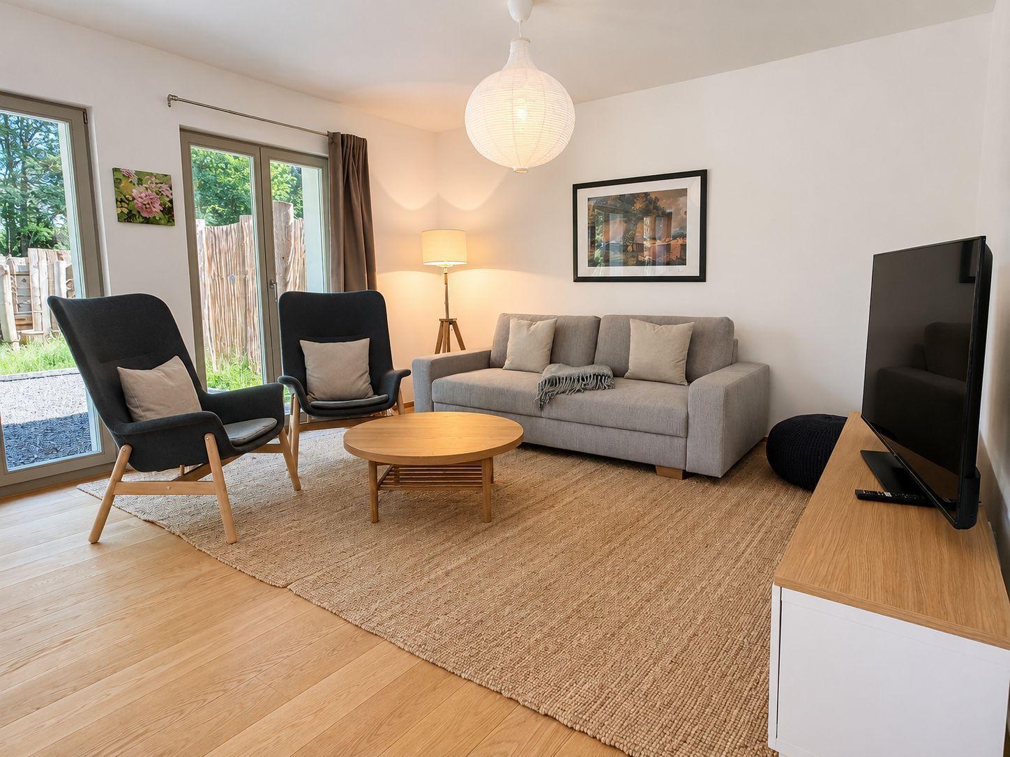 Wohnzimmer mit Sofa, zwei Sesseln, runden Tisch und Fernseher. Durchgangstür zum Garten.