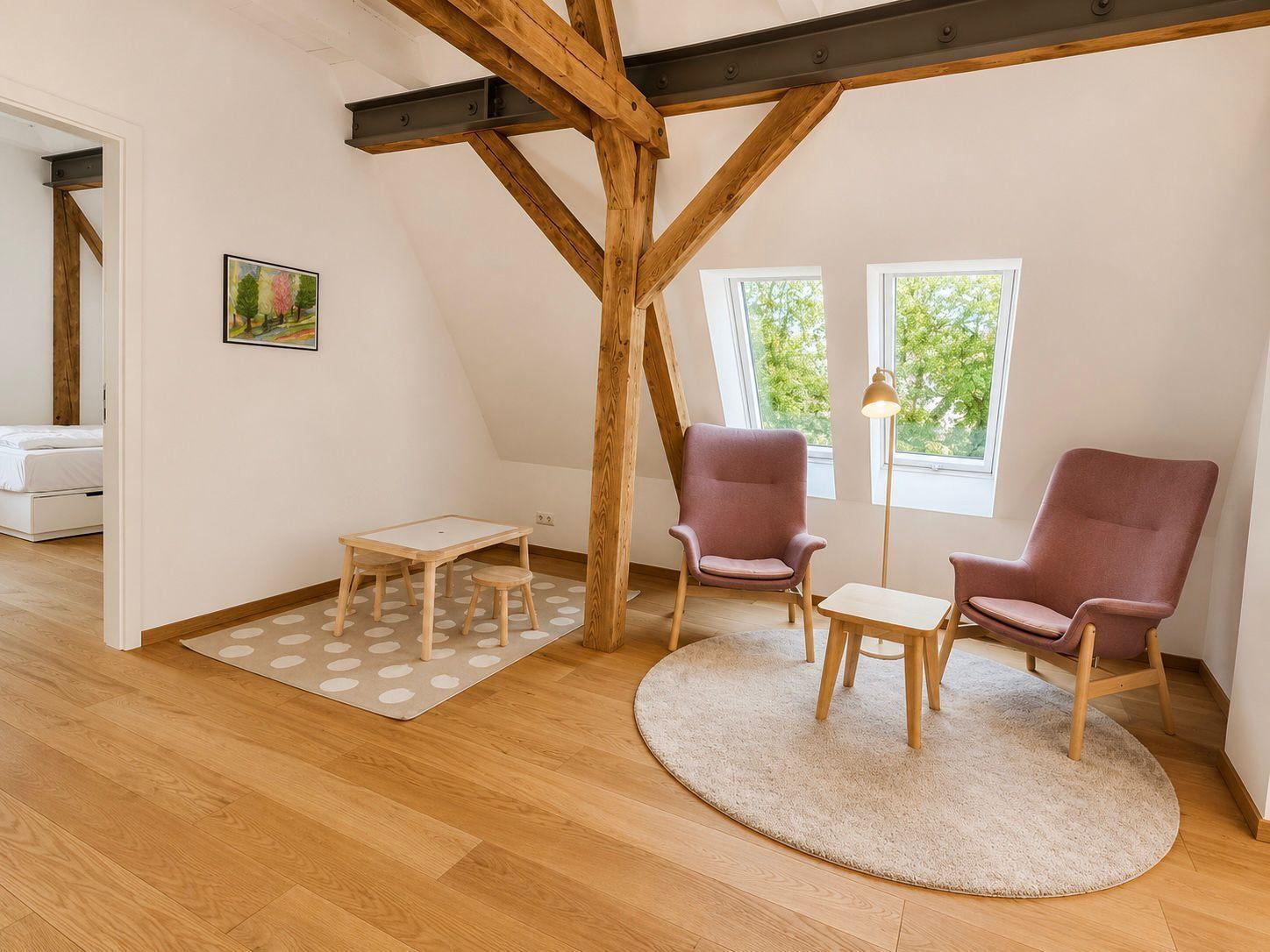 Zimmer mit Holzbalken, zwei Sesseln, Tisch und Fenstern. Im Hintergrund ist ein Schlafzimmer sichtbar.