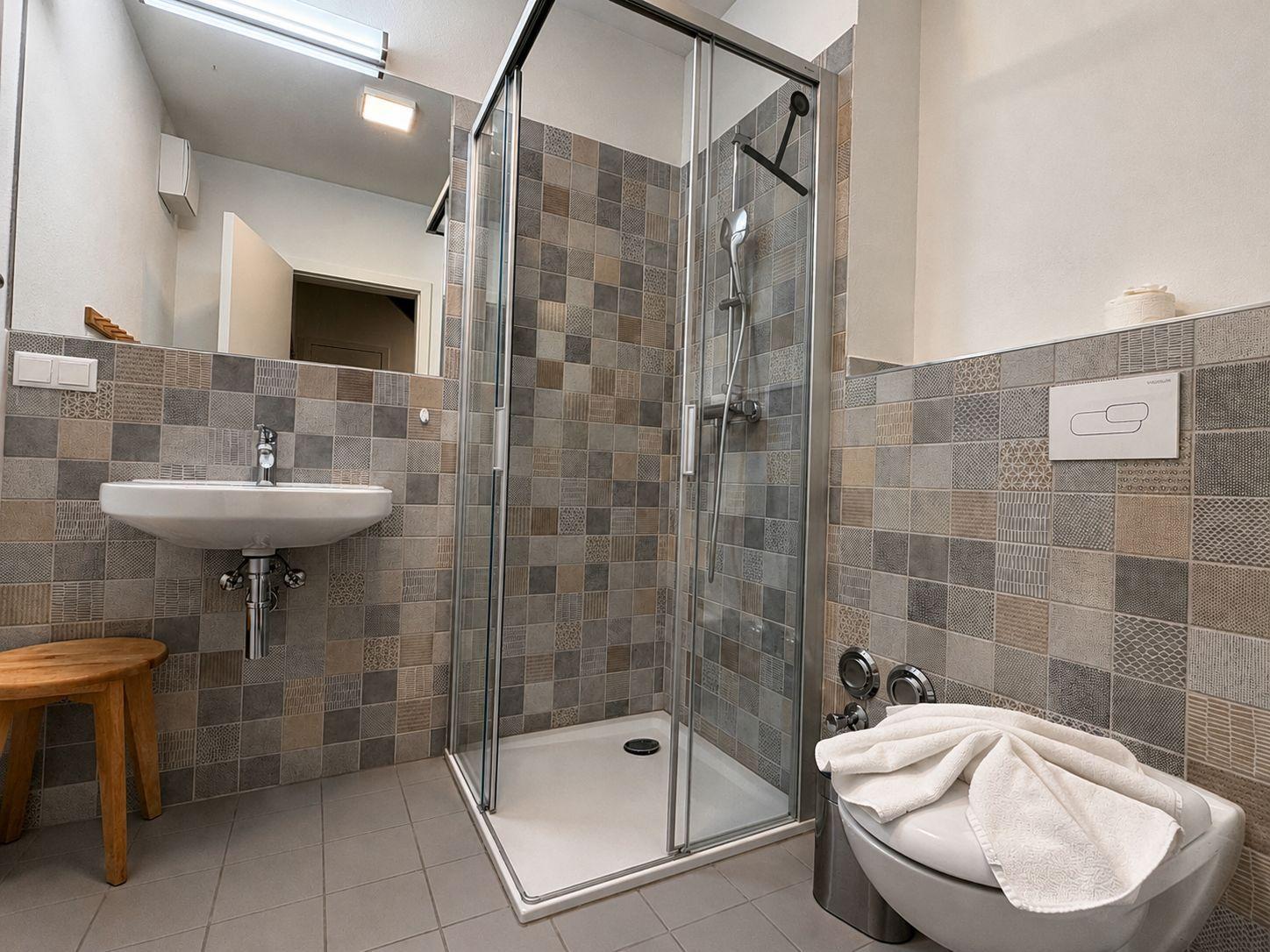 Badezimmer mit Dusche, Waschbecken und Toilette. Wandfliesen in verschiedenen Braun- und Grautönen.