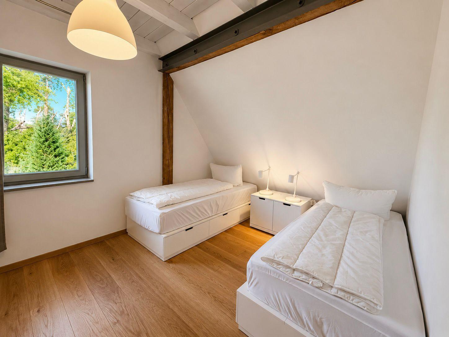 Zwei Betten mit weißen Laken, Holzboden und Fenster mit Blick auf Bäume.