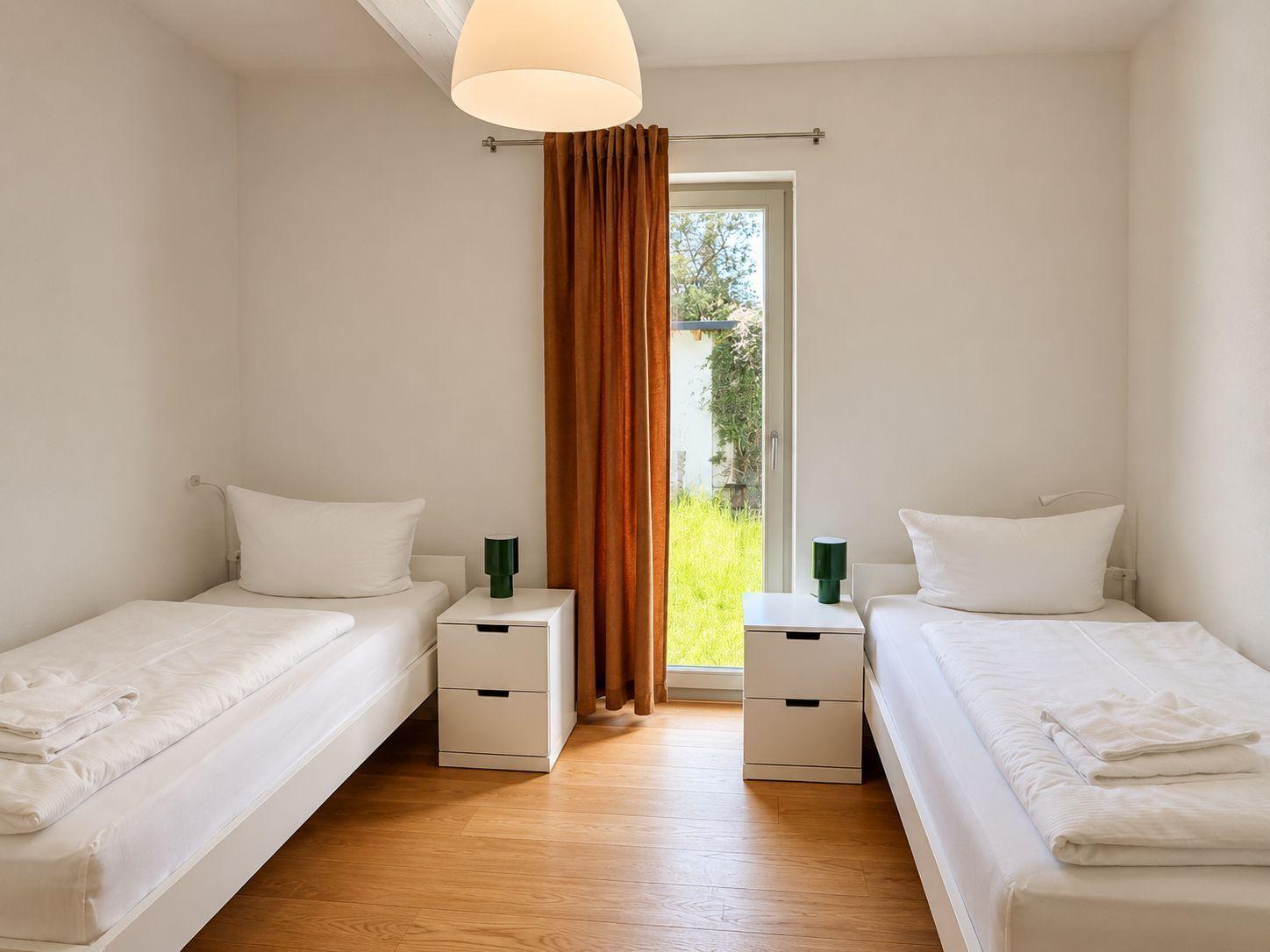 Zwei Einzelbetten mit Nachttischen und Fenster zur grünen Außenterrasse.