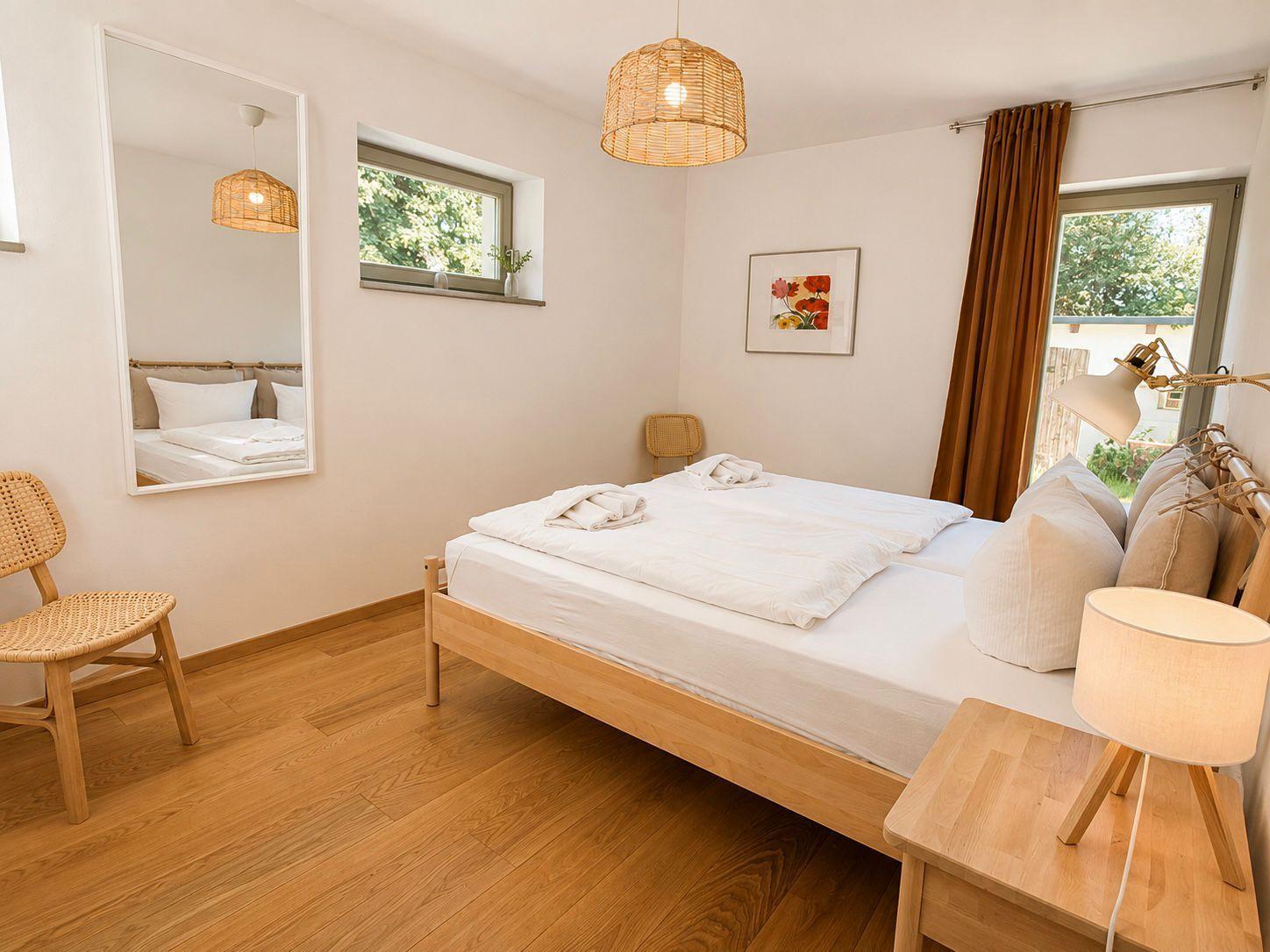 Schlafzimmer mit Holzbett, Spiegel, Lampe und Fenster mit Blick nach draußen.