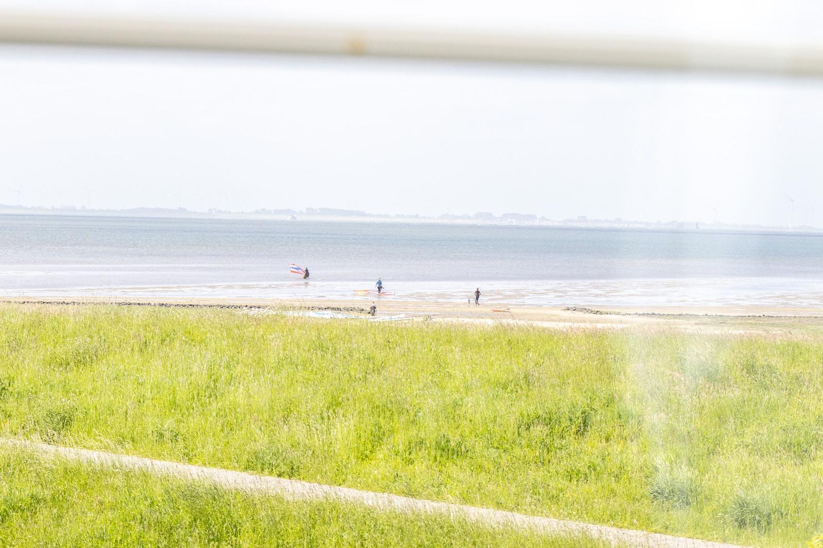 Grünes Gras am Ufer, Menschen im Wasser und Windräder im Hintergrund.
