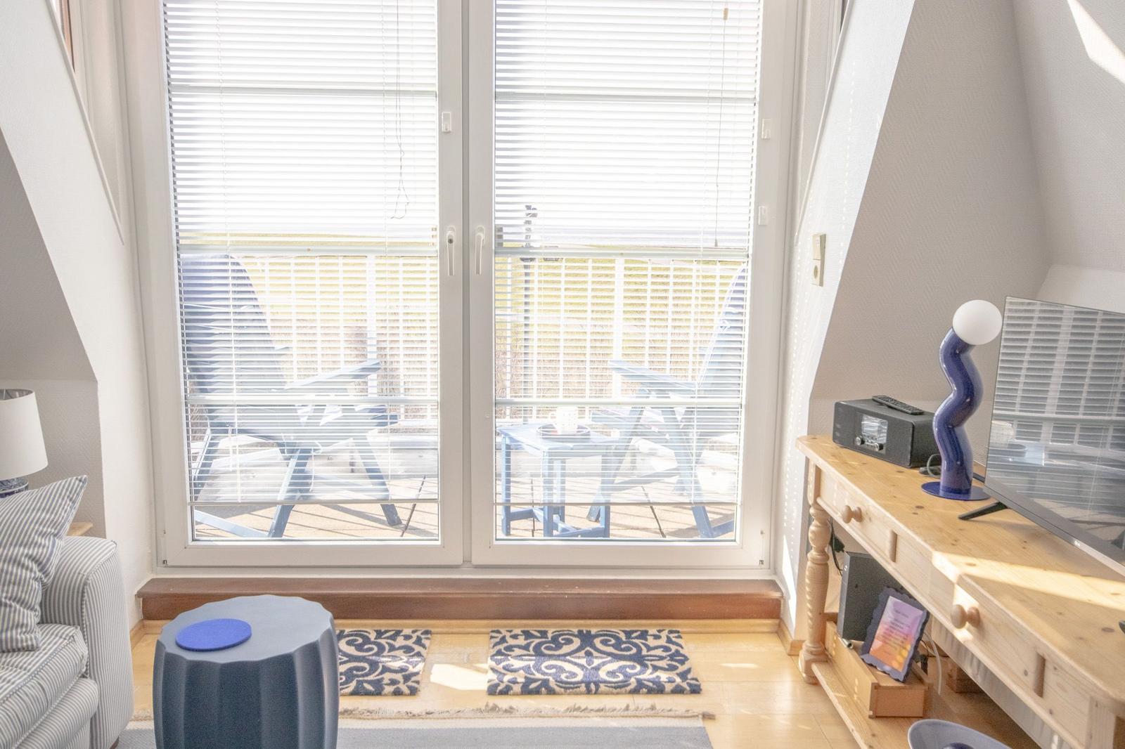 Durchgehendes Fenster mit Blick auf Balkon, Holzregal und Sitzgelegenheit im Wohnbereich.