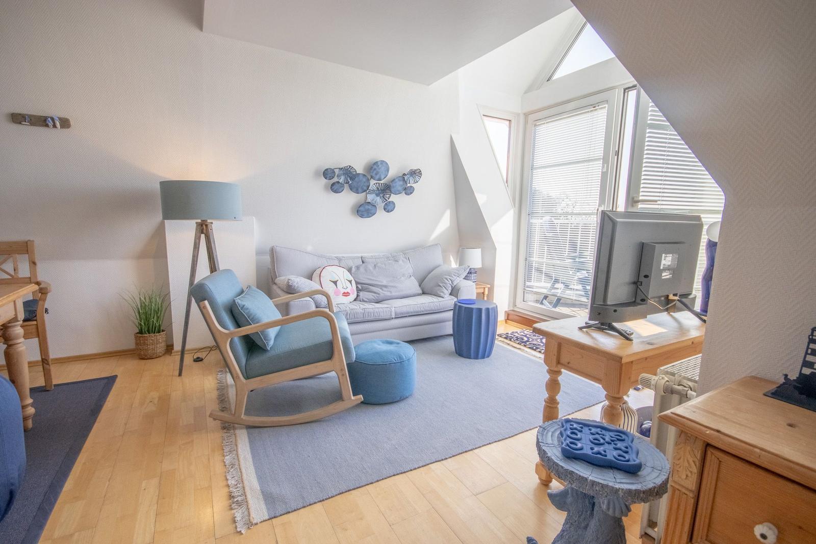 Wohnzimmer mit Sofa, Schaukelstuhl und TV. Holzboden und große Fenster.