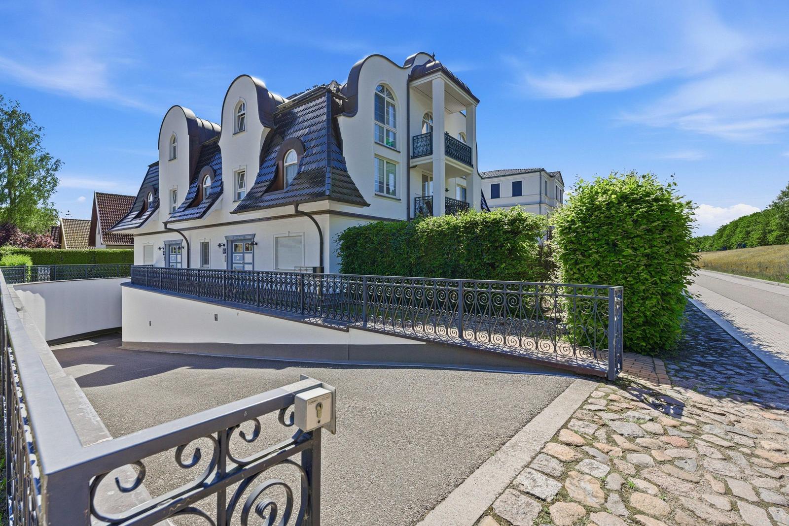 Einfahrt zu einer weißen Villa mit geschwungenen Dächern und schmiedeeisernem Zaun.