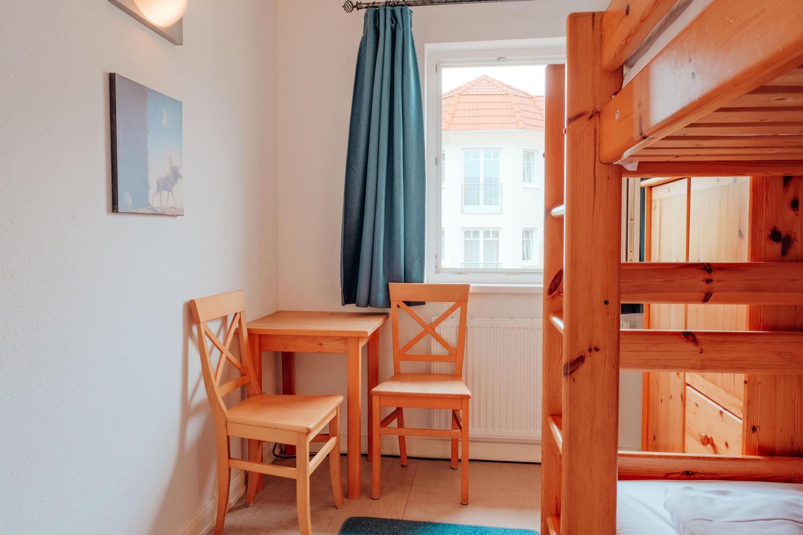 Zimmer mit Holzbett, Tisch und zwei Stühlen. Fenster mit Vorhang und Wandbild.