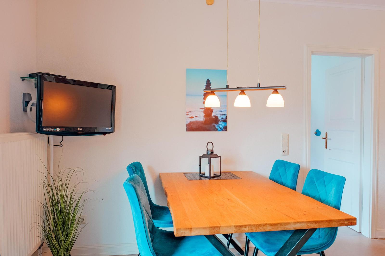 Esszimmer mit Holztisch, blauen Stühlen und Wandbild. An der Wand hängt ein Fernseher.