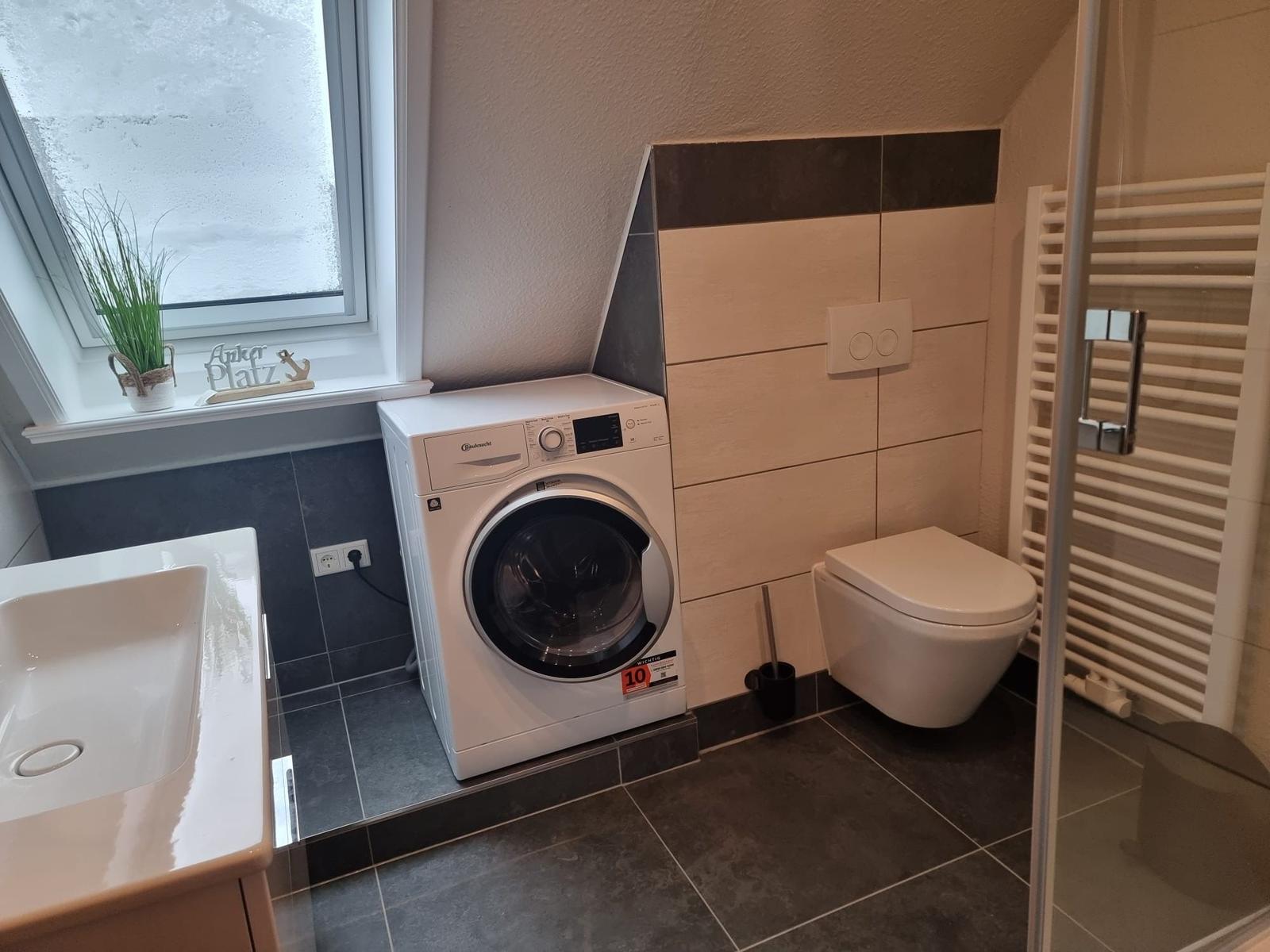 Bathroom with washing machine, toilet, and shower. Window with plant.