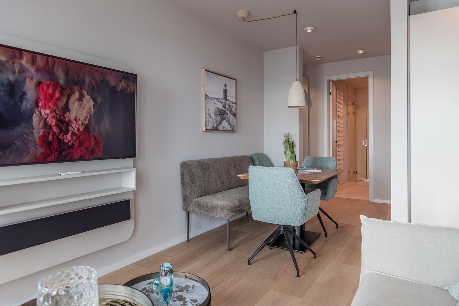 Wohn- und Essbereich mit Fernseher, Sitzbank und Stühlen. Holzboden und Wandbild sichtbar.