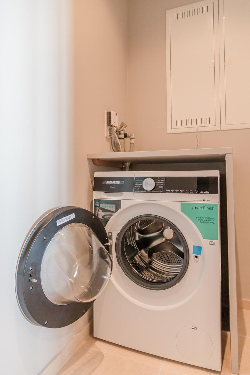 Waschmaschine mit offener Tür in einem Raum mit Schrank und Wasseranschluss.