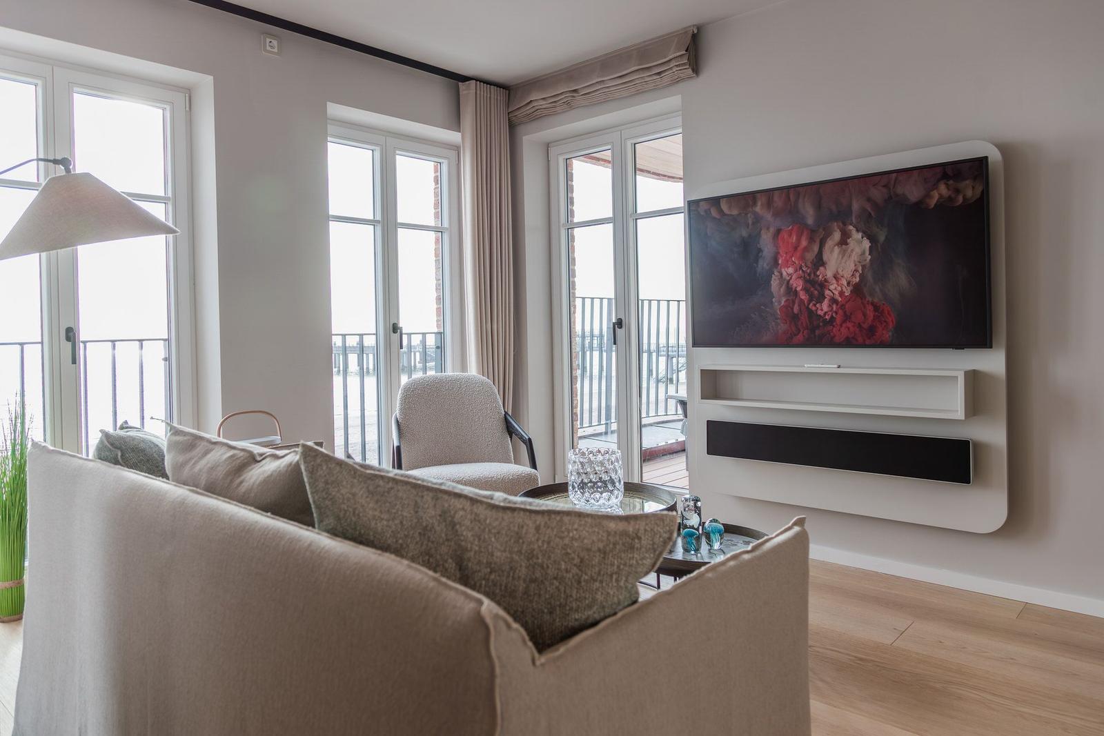 Wohnzimmer mit Sofa, Fernseher und Fenstern zum Balkon.