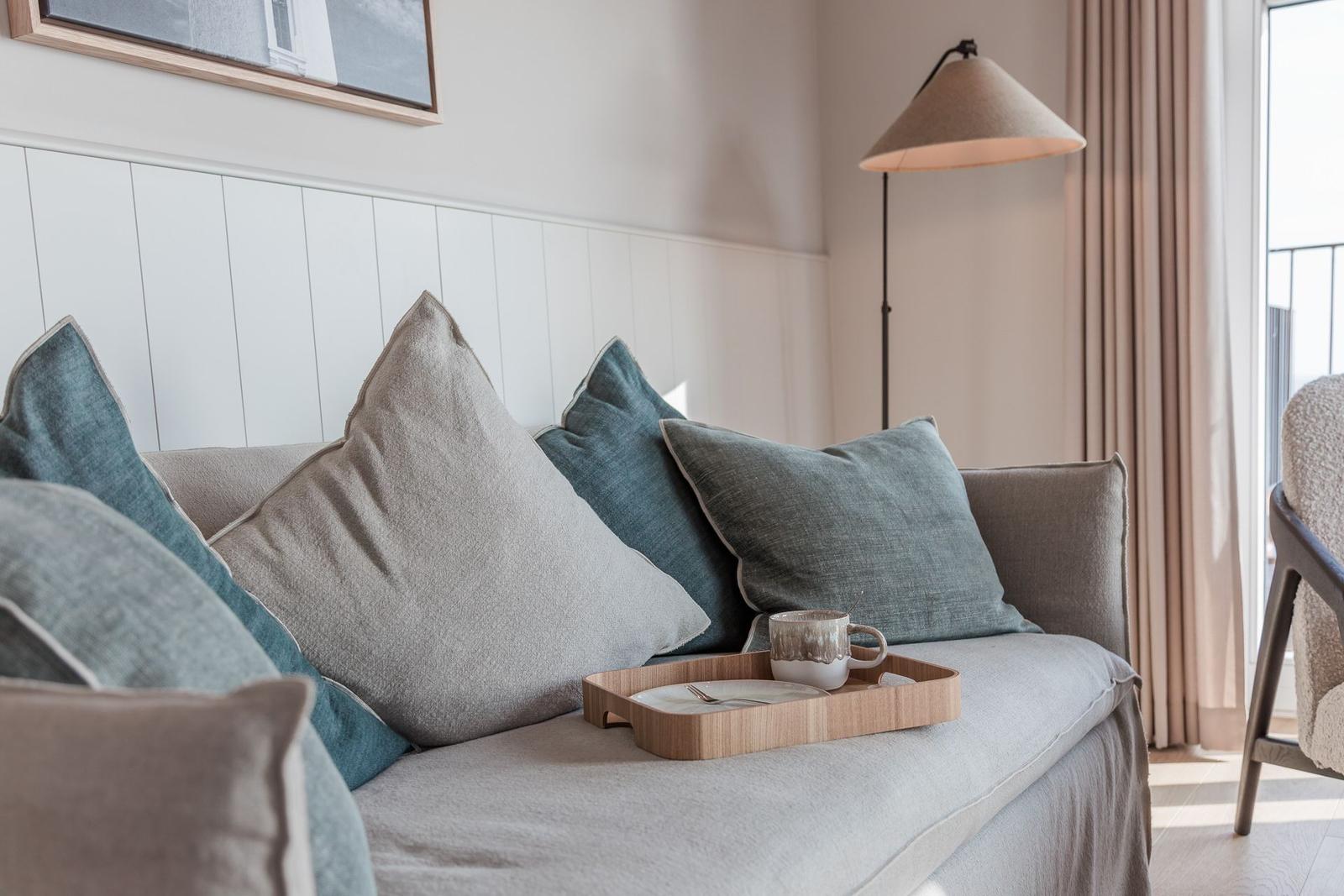 Ein Sofa mit Kissen und einem Tablett mit Tasse. Hintergrund: Wand mit Paneelen und Lampe.