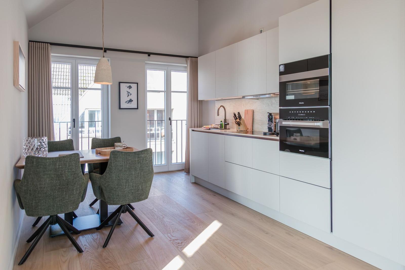 Küche mit Essbereich, weißen Schränken und integrierten Geräten. Holzfußboden und große Fenster.