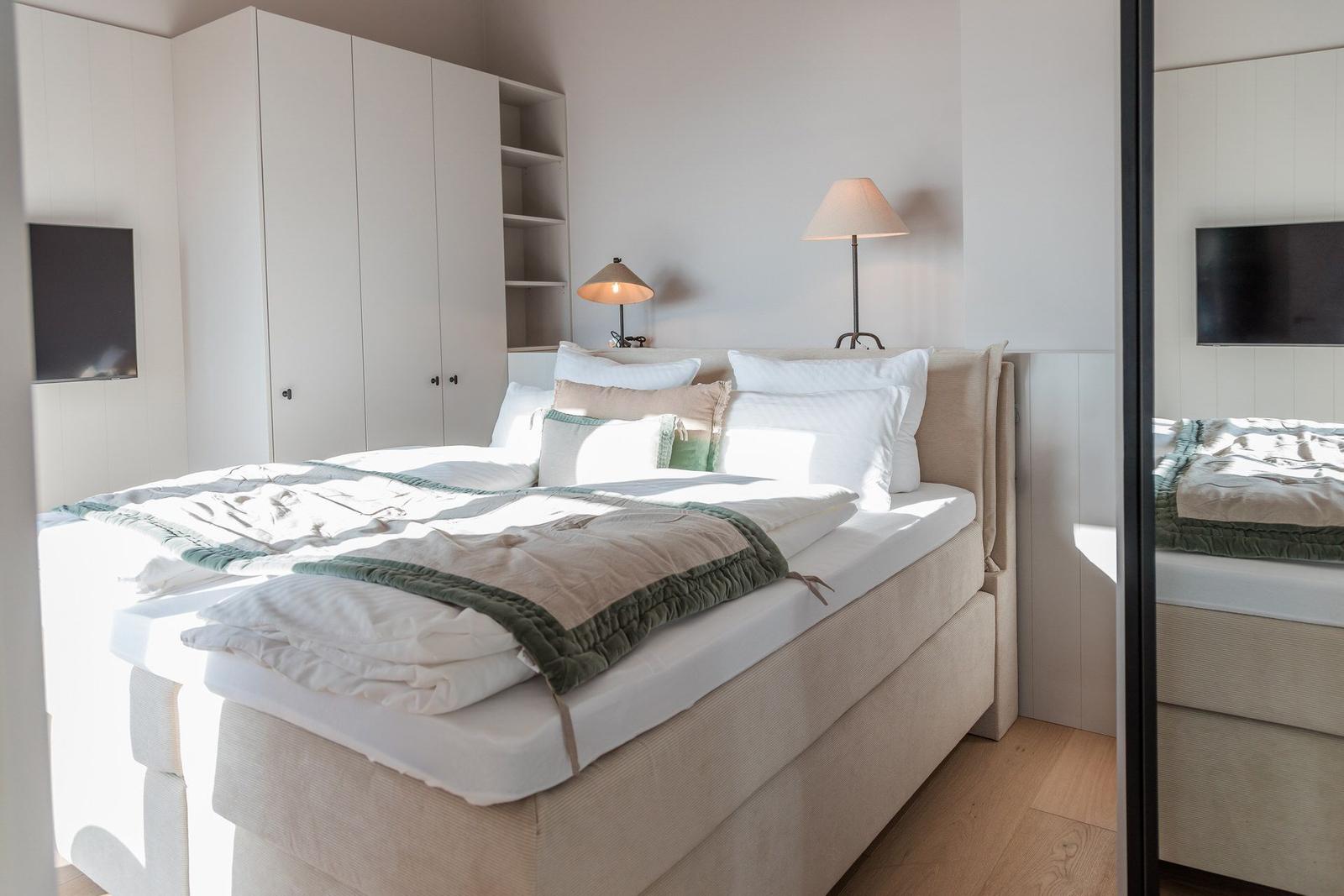 Schlafzimmer mit Bett, Nachttischlampen und Kleiderschrank. Spiegel reflektiert die Einrichtung.