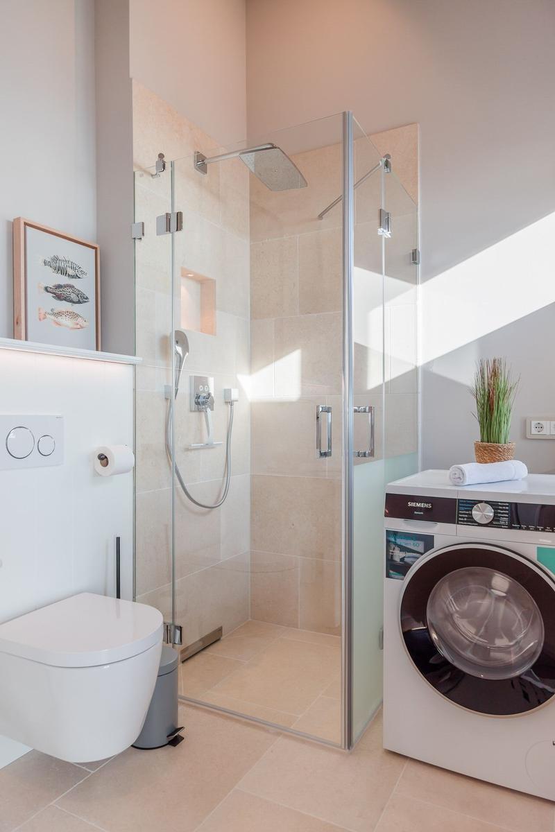 Dusche mit Glaswand, Waschmaschine und Toilette in einem hellen Raum.