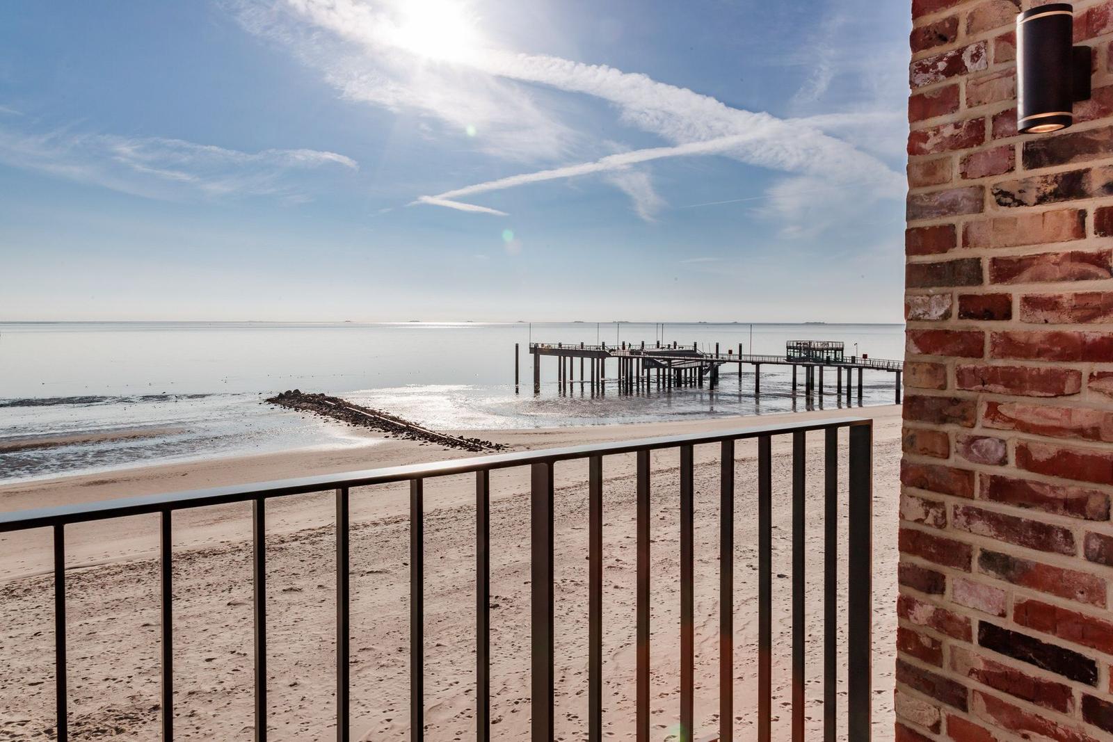 Blick von Balkon auf Strand und Pier unter blauem Himmel mit Flugzeugspuren.