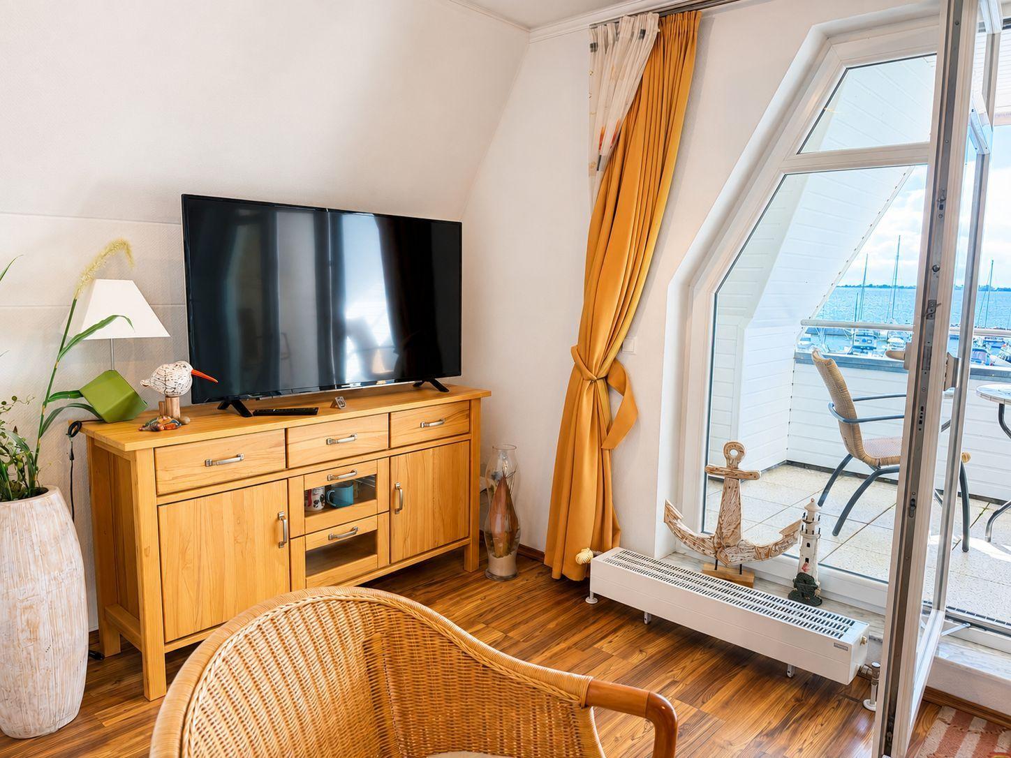 Ein Raum mit Fernseher auf Holzschrank, Balkon mit Blick auf Wasser und gelbe Vorhänge.