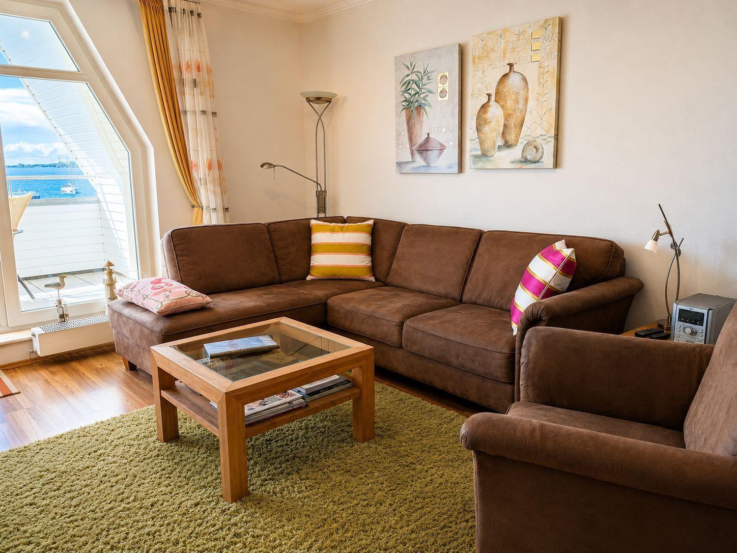 Wohnzimmer mit braunem Sofa, Glas-Tisch und Blick aufs Wasser.