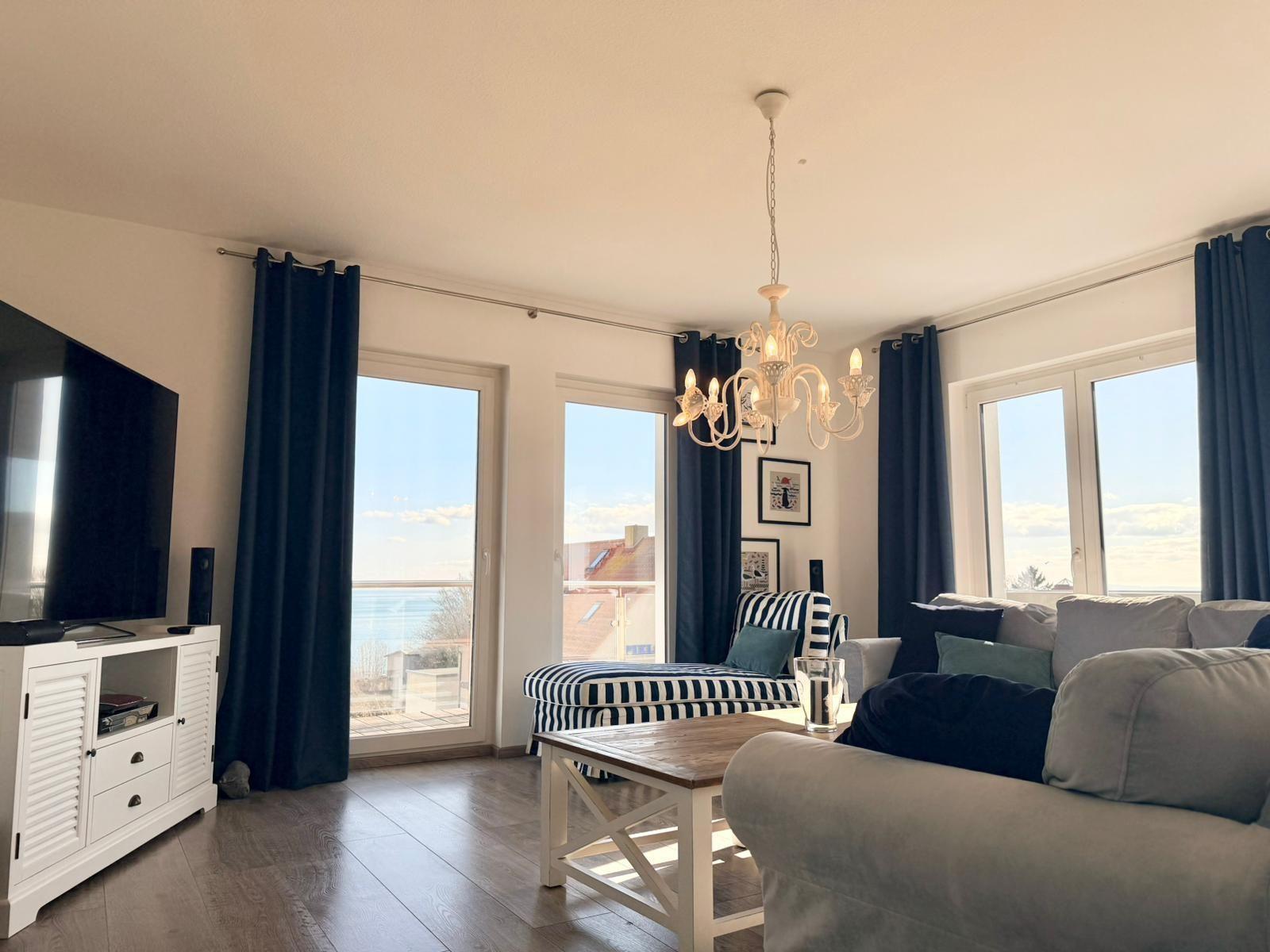 Wohnzimmer mit Sofa, Couchtisch und TV. Große Fenster mit Blick auf das Meer.