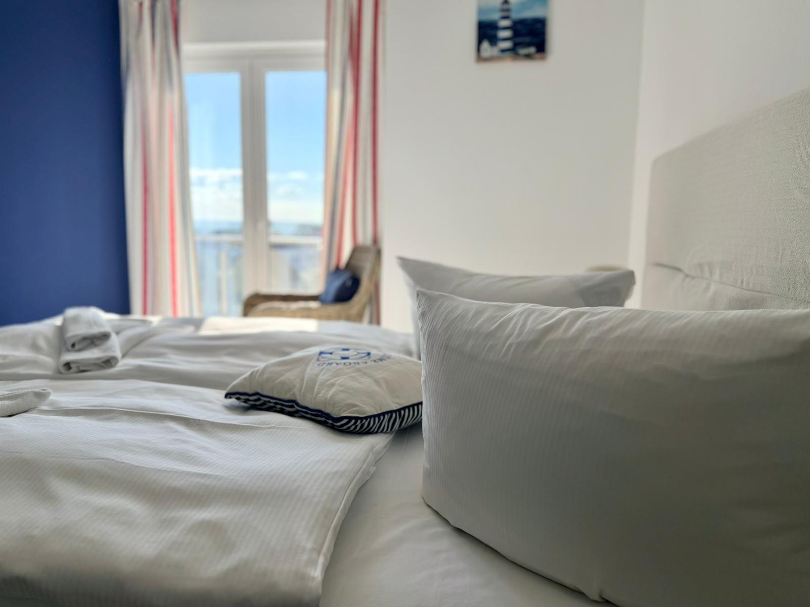 Ein Bett mit weißen Laken und Kissen. Im Hintergrund ein Fenster mit Blick auf Balkon und Meer.