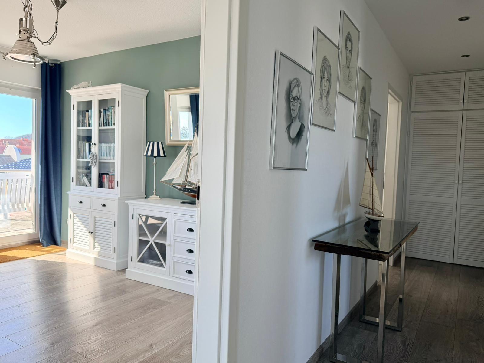 Hallenflur mit Bildern, Tisch und Modellboot. Links Wohnbereich mit Bücherregal und Balkon.