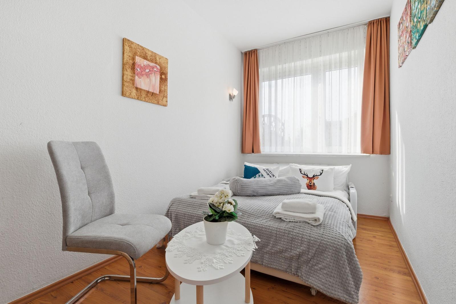 A bedroom with bed, chair and table. Window with curtains and wall art.
