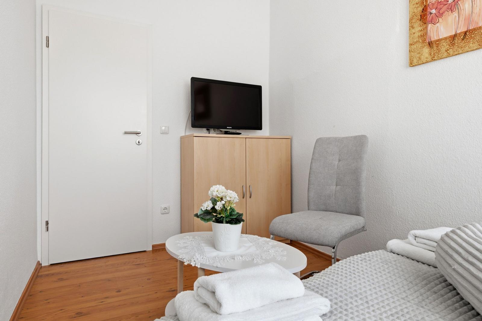 Room with bed, chair, TV and wooden floor. On table flowers and towels.
