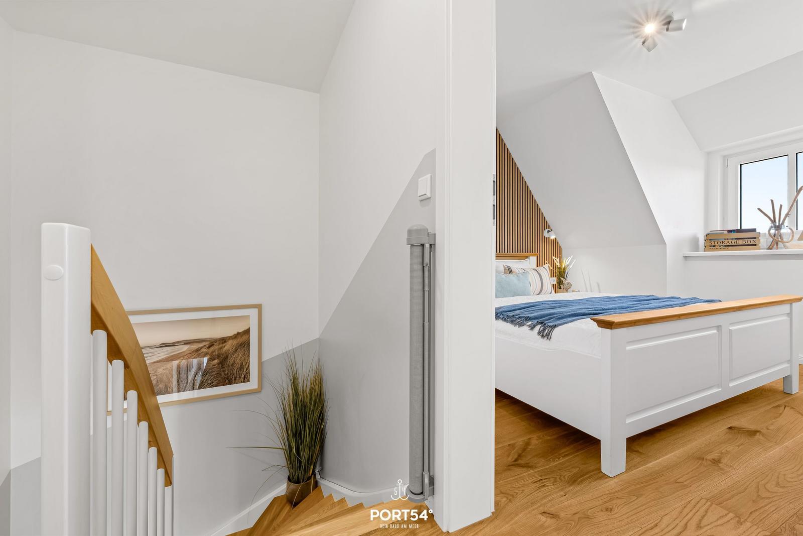 Master bedroom with sloped ceiling, white bed, and wooden floor. Staircase with wall art and plant.