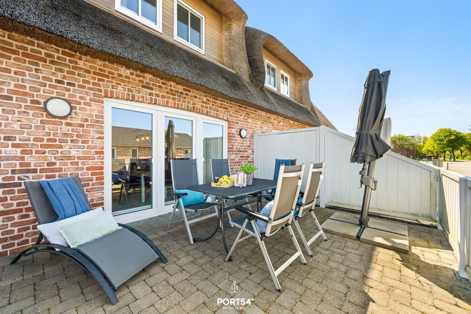 Terrace with table, chairs, and lounge chair in front of a house with brick facade and thatched roof.