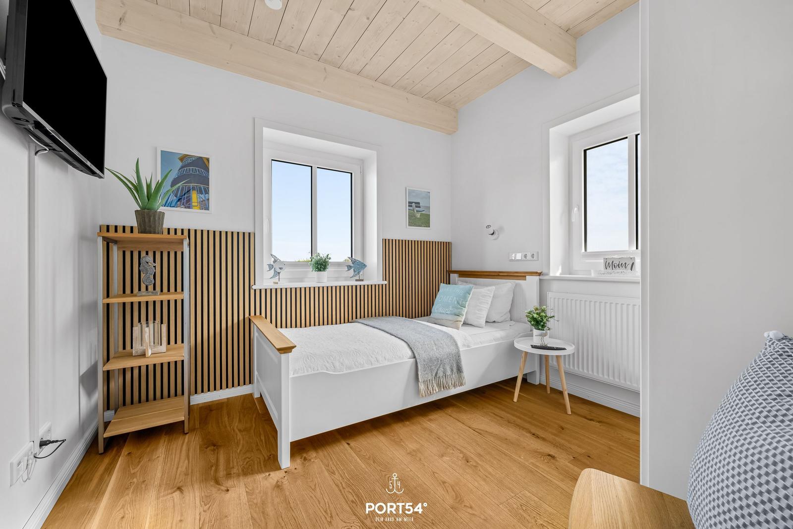 Bedroom with white bed, wooden floor, and two windows. Shelving unit and TV on the wall.