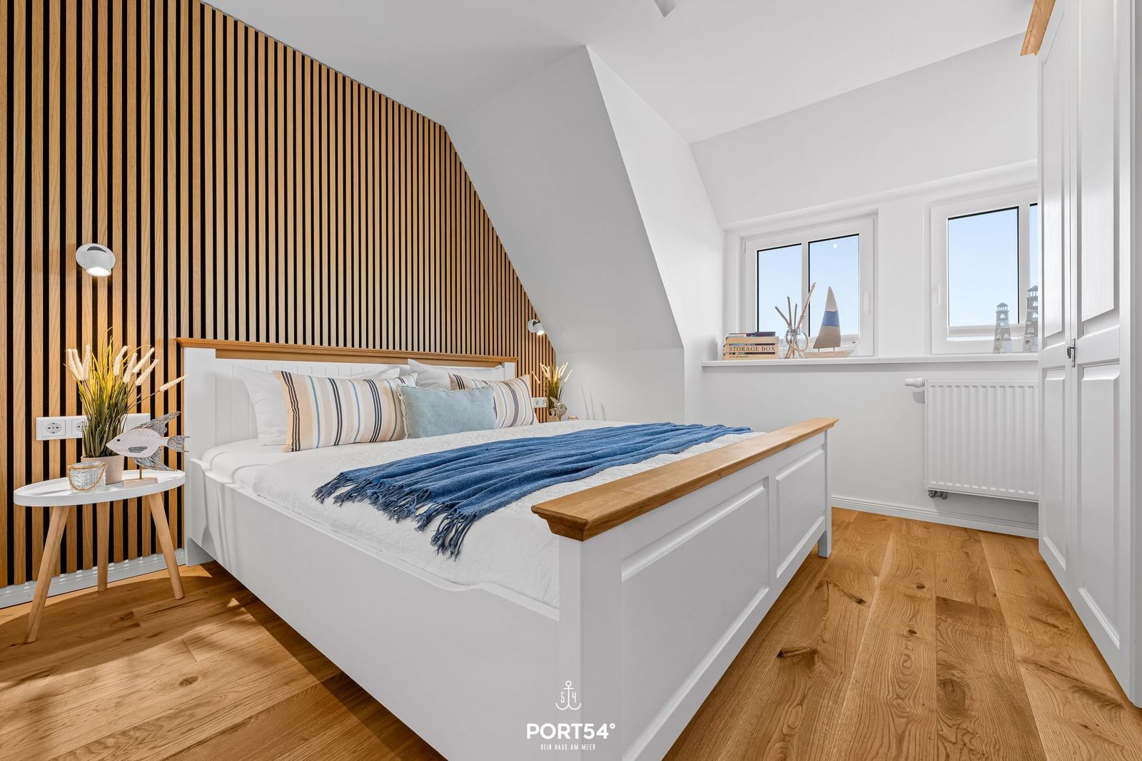 Schlafzimmer mit Holzdielen, weißem Bett und Holzwandverkleidung. Fenster mit Blick auf See.