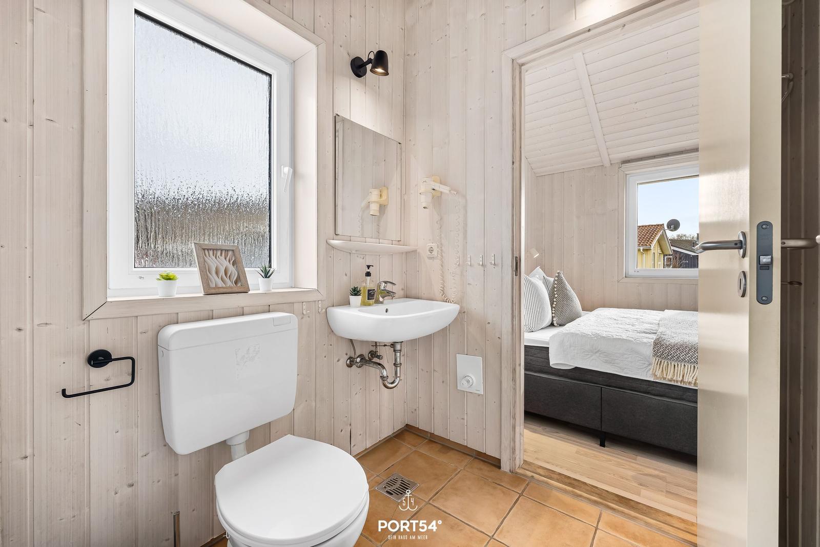 Badezimmer mit Toilette, Waschbecken und Fenster. Im Hintergrund Schlafzimmer mit Bett.