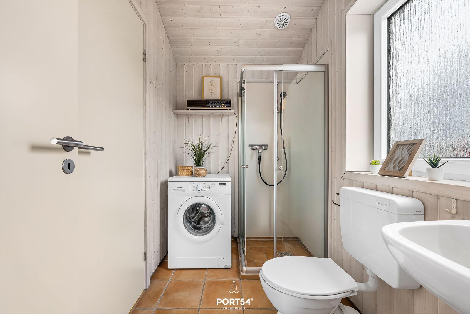 Badezimmer mit Dusche, Waschmaschine und Toilette. Holzwand und Fenster mit Glas.
