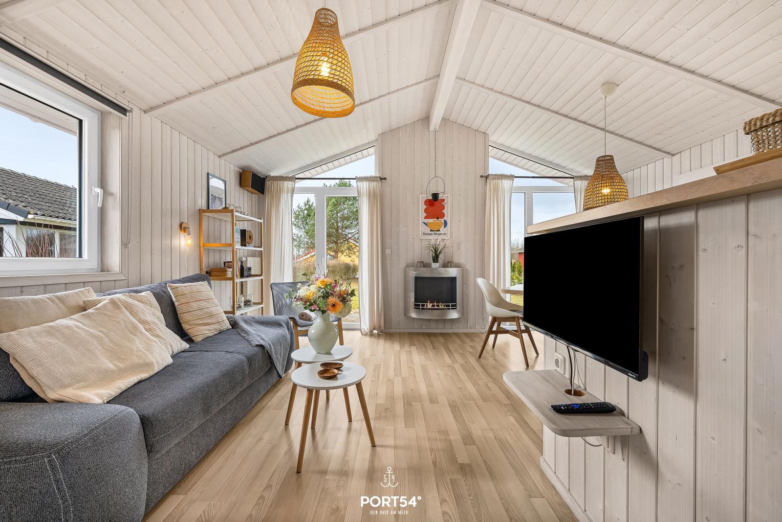 Wohnraum mit Sofa, Fernseher und Kamin. Holzboden und große Fenster.