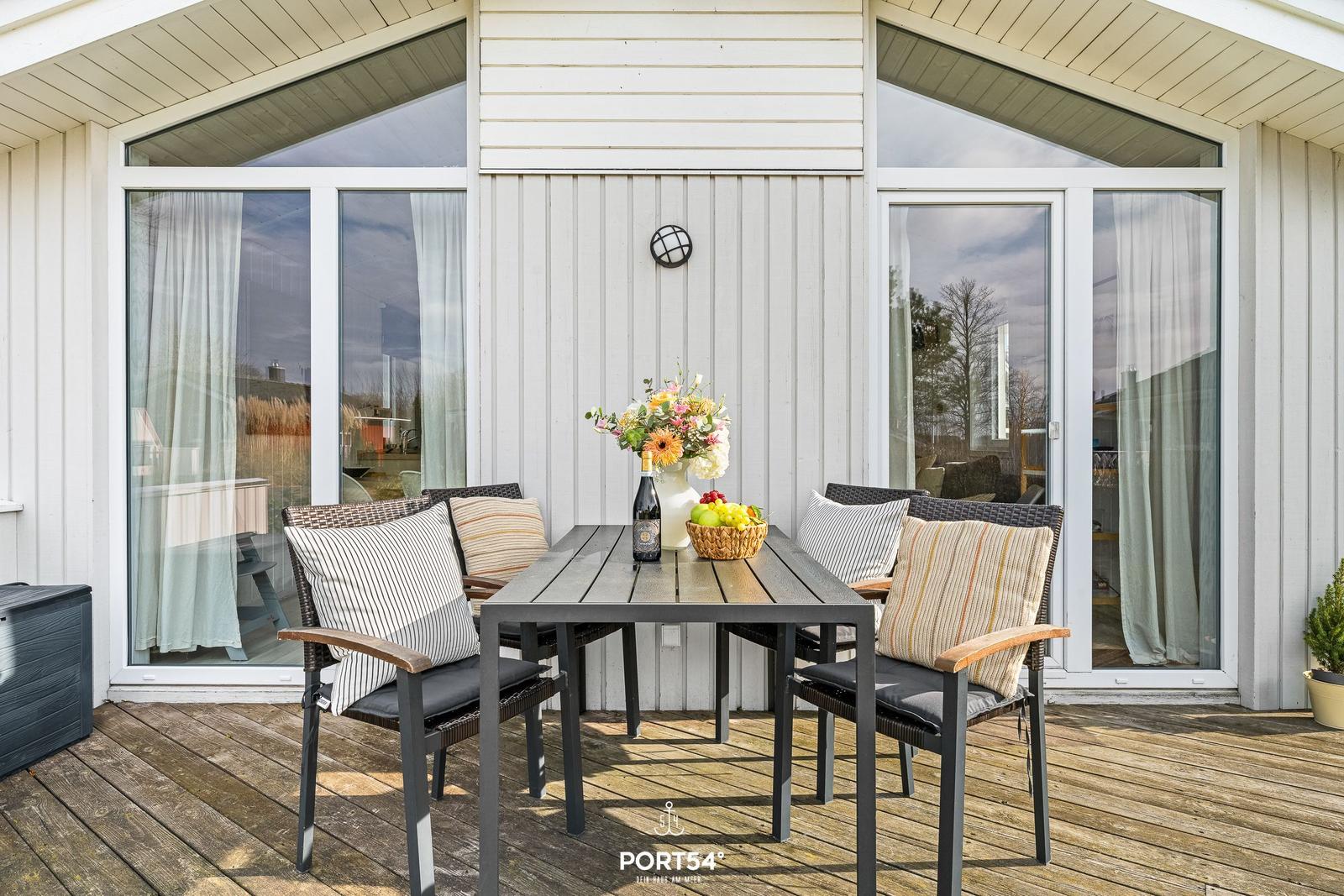 Terrasse mit Tisch, Stühlen und Blumen. Holzdecke und große Fenster.